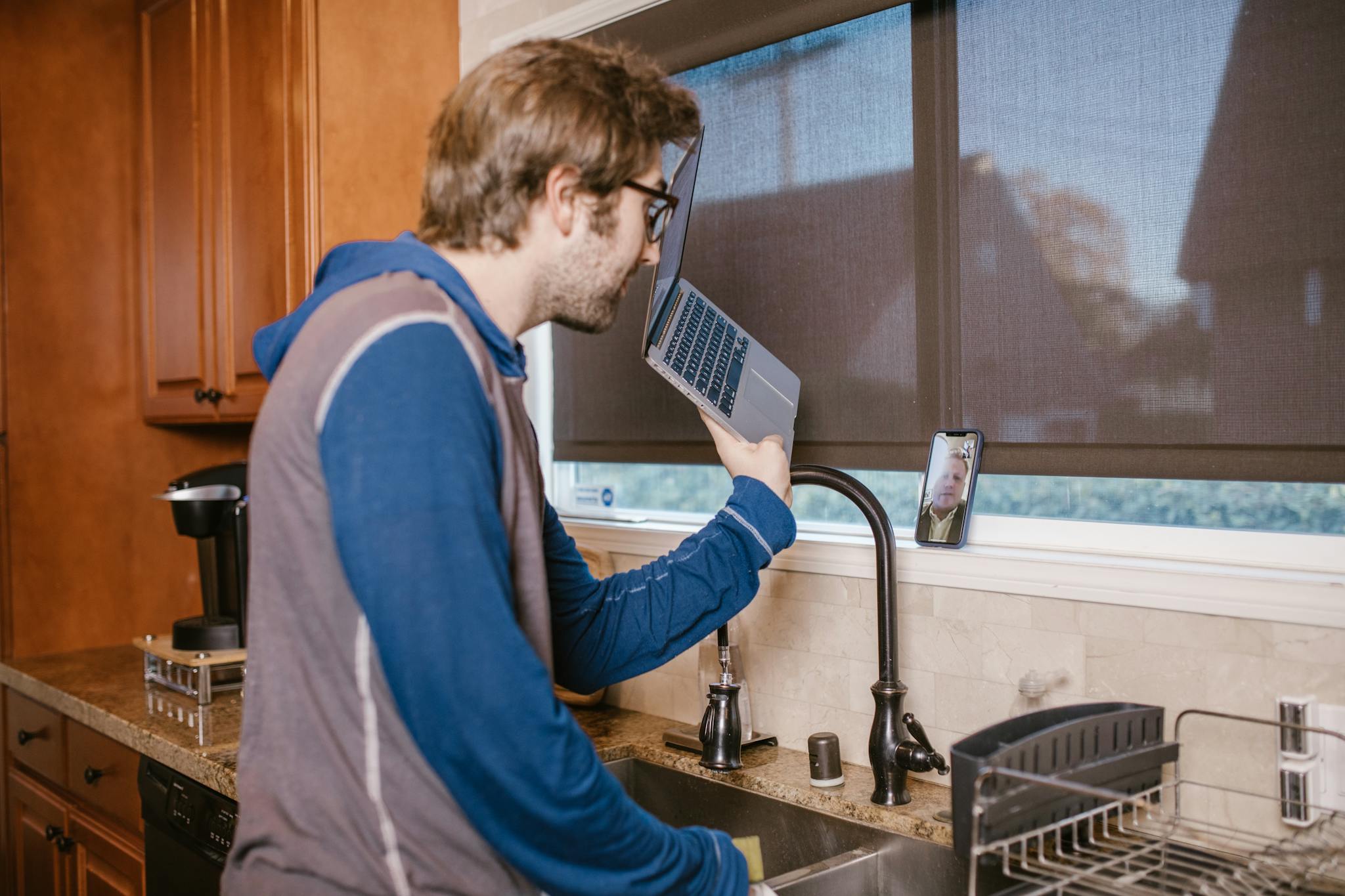A man in a kitchen making a video call using a laptop and smartphone.